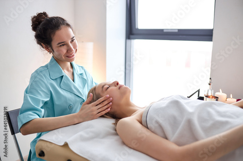 woman receiving osteopathic or chiropractic treatment on face and head in clinic, side view on female lying on bed getting massage procedure by woman, relaxing with eyes closed, side view portrait photo