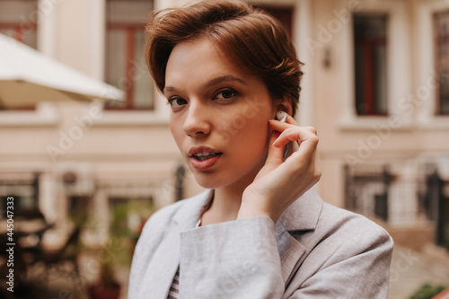 Lady sensitively looks into camera and posing outside. Close-up portrait of short-haired stylish pretty woman dressed in grey jacket. Girl wearing wireless headphones