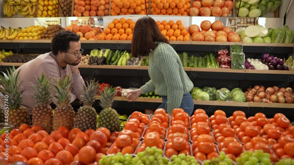 Healthy eating woman choosing vegetarian products in grocery store while bored mixed race boyfriend watching her leaning on shopping cart. Multiracial couple of buyers making purchases in farm shop Healthy eating woman choosing vegetarian products in grocery store while bored mixed race boyfriend watching her leaning on shopping cart. Multiracial couple of buyers making purchases in farm shop