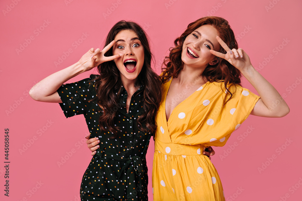 Curly ladies having fun on pink background and showing peace signs. Beautiful women in polka dot outfits are smiling beautifully and sincerely Curly ladies having fun on pink background and showing peace signs. Beautiful women in polka dot outfits are smiling beautifully and sincerely