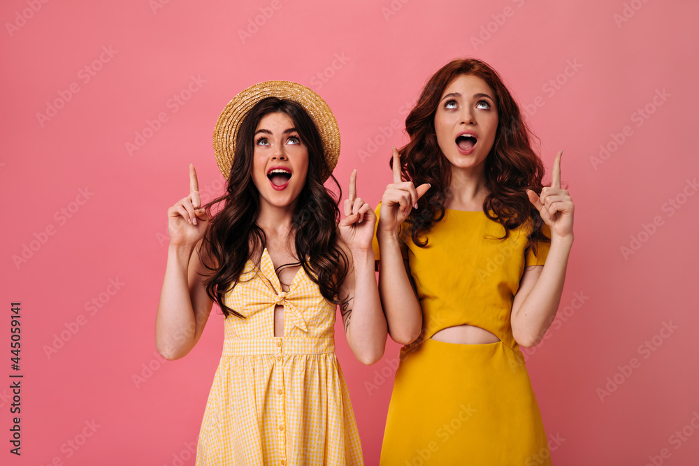 Women in yellow outfits pointing up on pink background. Modern lady with wavy hair poses with her fashionable girlfriend in summer dress