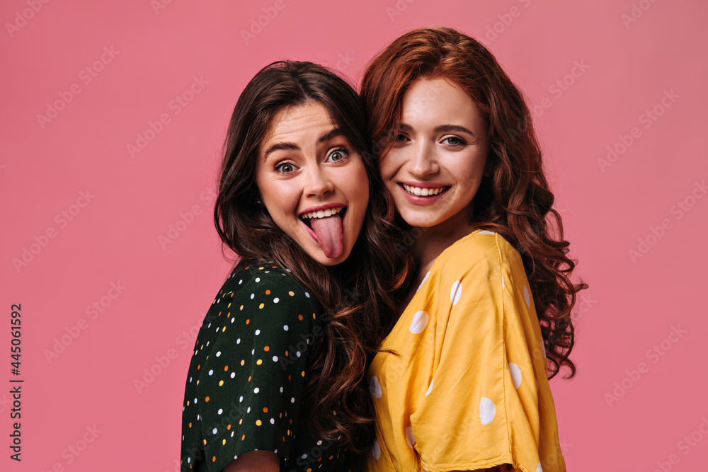 Mischievous Girl in black dress demonstrates her tongue. Ginger woman in bright yellow blouse cute smiling. Two curly ladies posing on pink background