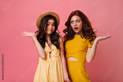 Shy Girls in yellow dresses pose on pink background. Lovely ladies with wavy dark hair in summer clothes posing for camera