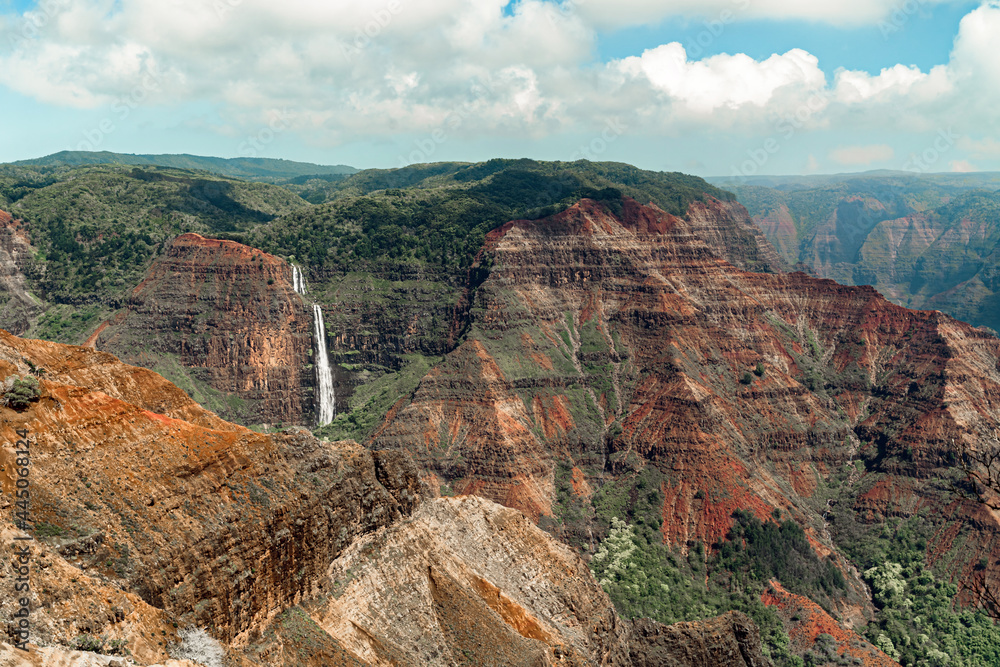 A small waterfall is almost eclipsed by the dramatic red rock of the Hawaiian Waimea Canyon. Called the Grand Canyon of the Hawaiian Islands, it is one of the most visited national parks  in Kawaii