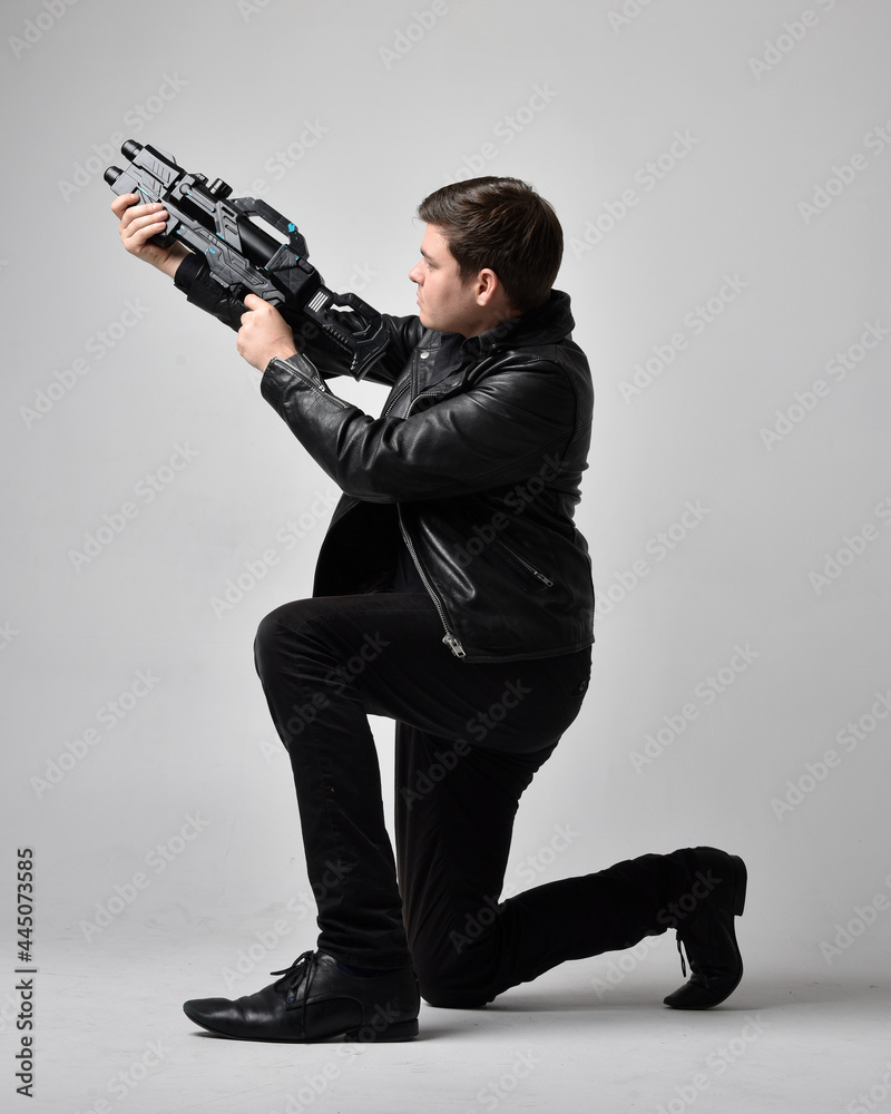 Full length portrait of a brunette man wearing leather jacket and holding a science fiction gun. kneeling action pose isolated against a grey studio background.
Full length portrait of a brunette man wearing leather jacket and holding a science fiction gun. kneeling action pose isolated against a grey studio background.