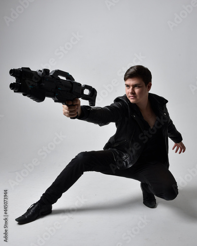 Full length portrait of a  brunette man wearing leather jacket  and holding a science fiction gun.  kneeling  action pose isolated  against a grey studio background.
