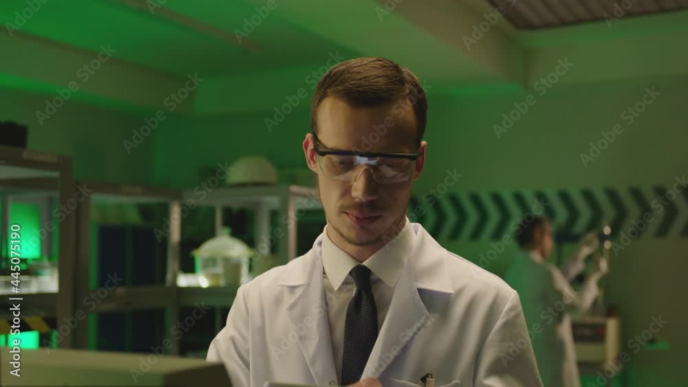 Young male scientist on white lab coat working on laboratory , using tablet and doing notes . Men working inside high tech lab room Laboratory equipment and lab shelves made special for this footage Young male scientist on white lab coat working on laboratory , using tablet and doing notes . Men working inside high tech lab room Laboratory equipment and lab shelves made special for this footage