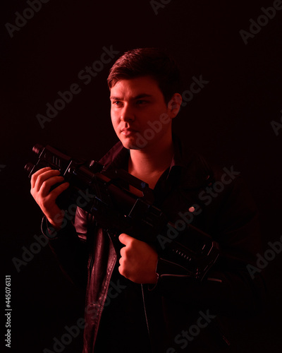  close up portrait of a  brunette man wearing leather jacket  and holding a science fiction gun.  Standing  action pose with red silhouetted lighting against a black studio background.