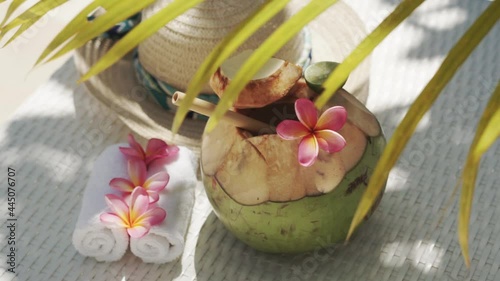Video footage of green young coconut close up with bamboo straw, sun hat, tropical flowers frangipani, palm shade on white sun bed in Bali 