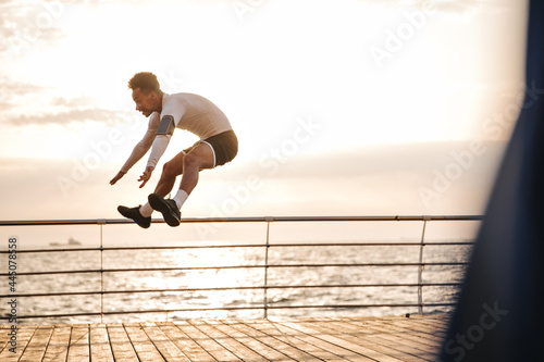 Portrait of young brunette man in black sport shorts and white t-shirt jumping high near sea during sunset.
