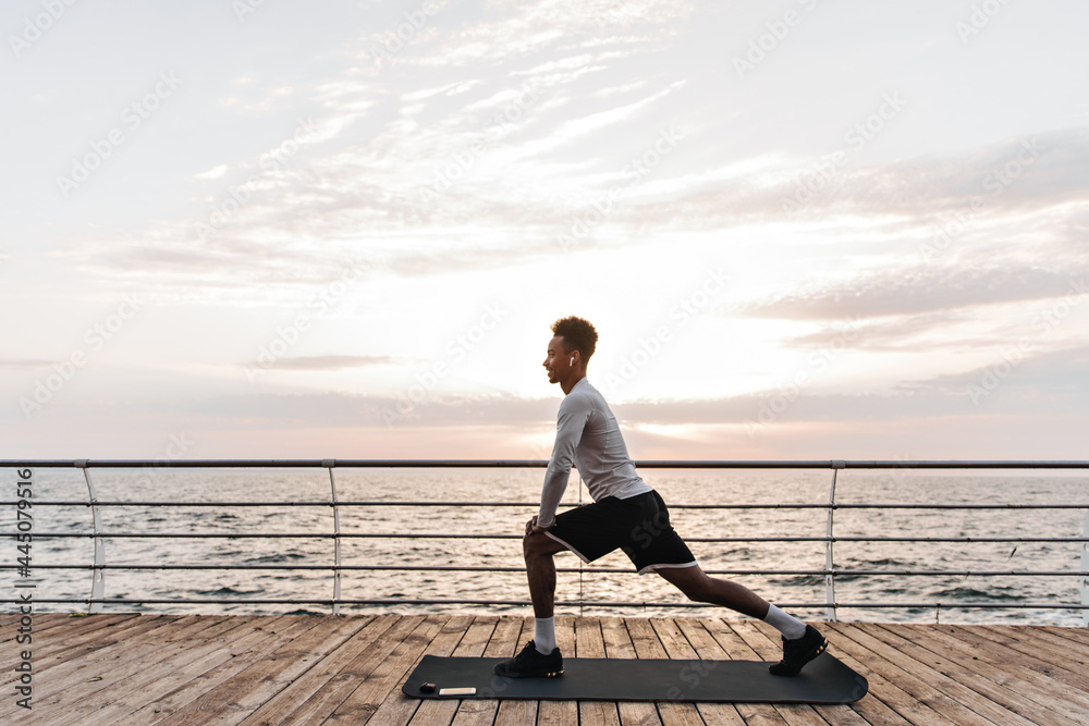 Handsome curly dark-skinned man in black shorts and white t-shirt stretches near sea. Cool guy poses on yoga mat outside.