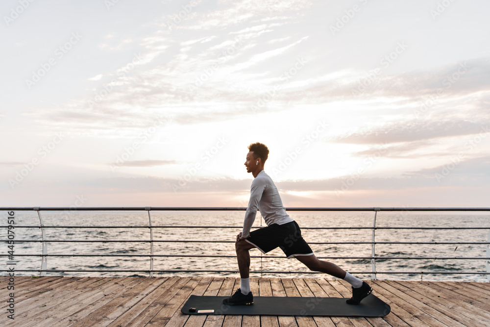 Handsome curly dark-skinned man in black shorts and white t-shirt stretches near sea. Cool guy poses on yoga mat outside. Handsome curly dark-skinned man in black shorts and white t-shirt stretches near sea. Cool guy poses on yoga mat outside.