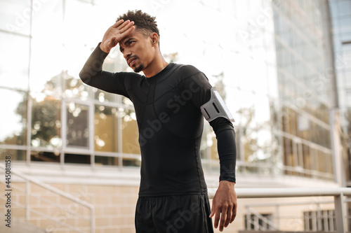 Handsome curly dark-skinned man in black long-sleeved t-shirt and shorts touches his face. Hardworking sportsman working out.