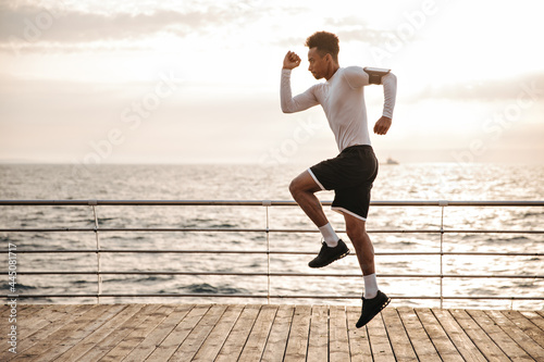 Strong young curly dark-skinned man in black sport shorts and long-sleeved t-shirt jumps and runs outside near sea.