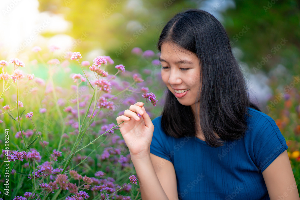 Portrait of a beautiful long-haired Asian woman in a blue shirt looking happily at the purple flowers in the garden with a smiling face.