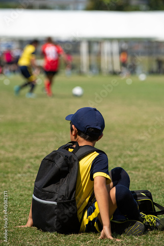 Junior soccer player rests with his teammates on a sideline waiting for their next match in a football school tournament, while some pitches still in action.