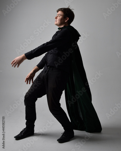 Full length portrait of a brunette man wearing black shirt, waistcoat and a green velvet cloak. Standing action pose isolated against a grey studio background.