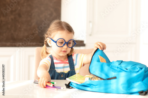 Girl in glasses takes out school stationery from his backpack to do homework at table on sunny morning kitchen.Education,learning.Back to school.Place for text, mock up