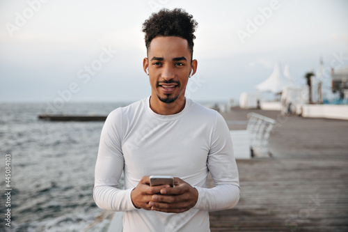 Calm serious dark-skinned man in white long-sleeved t-shirt looks into camera, holds phone and listens to music in headphones.