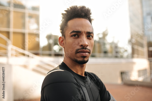 Close-up portrait of brunet young dark-skinned man in sport black t-shirt looks straight. Serious guy poses outside.