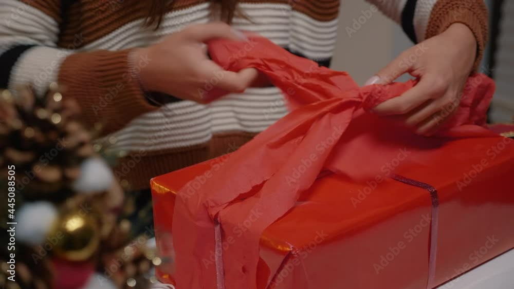 Young adult wrapping gift box in red paper to give to friends at christmas eve celebration party. Woman decorating present with ribbon for winter season joyful tradition in december