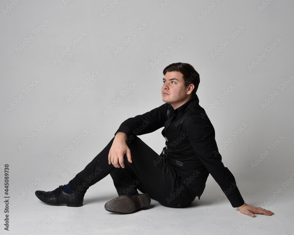 Full length portrait of a brunette man wearing black shirt and gothic waistcoat. Seated pose isolated against a grey studio background. Full length portrait of a brunette man wearing black shirt and gothic waistcoat. Seated pose isolated against a grey studio background.