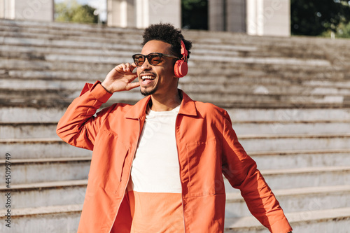 Good-humored curly brunet man in red headphones listens to music and sings. Charming guy in sunglasses poses outside near stairs.