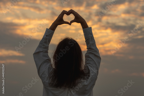 Young Woman holding a heart shape on the ocean, sea shore sunset evening.Holiday concept.Back view.