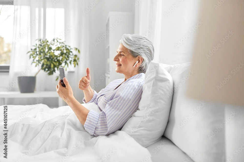 technology, old age and people concept - senior woman with smartphone and wireless earphones having video call in bed at home bedroom technology, old age and people concept - senior woman with smartphone and wireless earphones having video call in bed at home bedroom