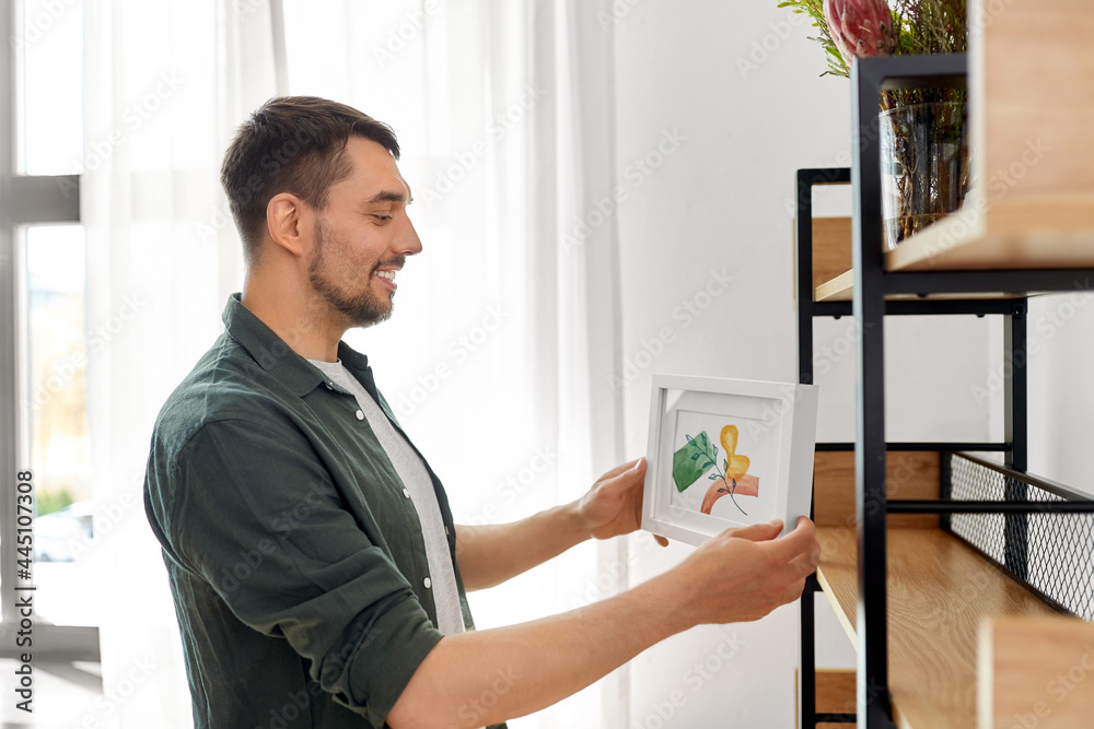 home improvement, decoration and people concept - happy smiling man placing watercolor picture in frame to shelf home improvement, decoration and people concept - happy smiling man placing watercolor picture in frame to shelf