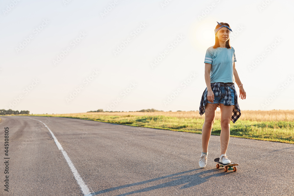 Beautiful female wearing t shirt, short and hair band skateboarding in the street, looking in distance, spending time alone with pleasure, active and healthy lifestyle.