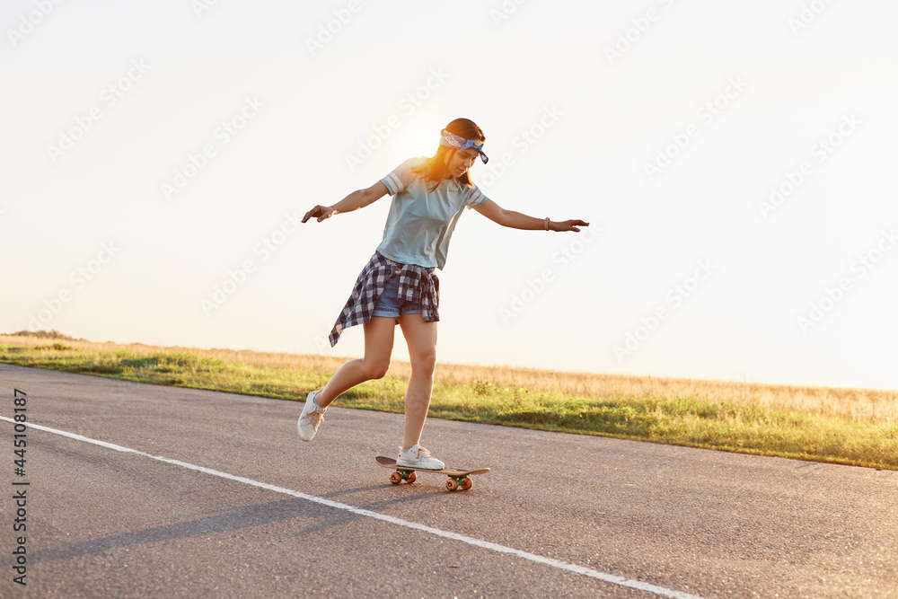 Fototapeta premium Young attractive female skater riding outdoor on asphalt road, raised her arms, sporty woman wearing casual clothing skateboarding alone in sunset in summer.