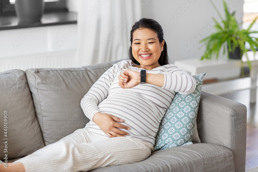 pregnancy, rest, people and expectation concept - happy smiling pregnant asian woman with smart watch sitting on sofa at home