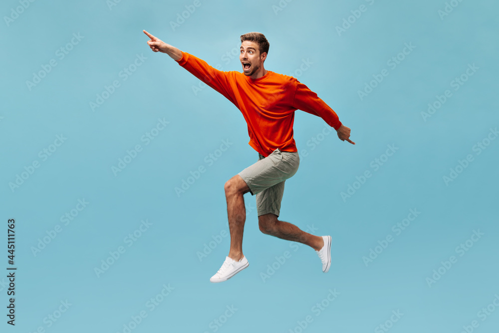 Positive guy with beard in bright cool clothes and white modern sneakers looking away and jumping on isolated blue background. .