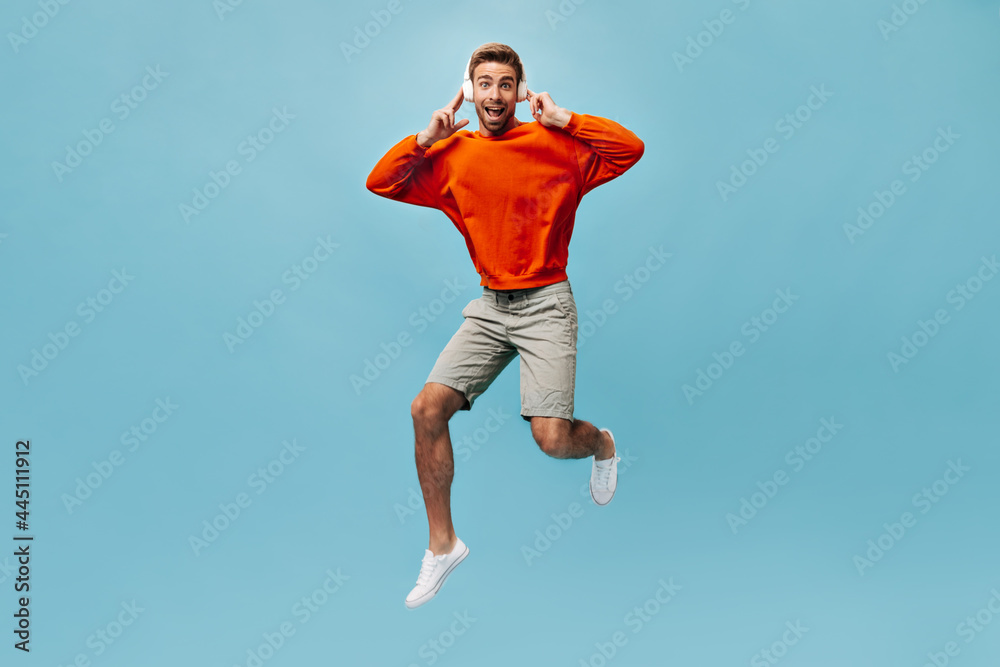 Fototapeta premium Handsome guy in good mood in orange sweatshirt, shorts and white sneakers looking into camera, listening to music and jumping..