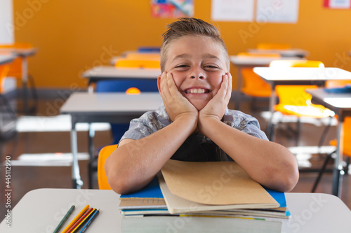 Happy caucasian schoolboy sitting at desk in classroom leaning on books and smiling