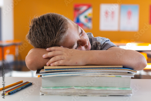 Tired caucasian schoolboy sitting at desk in classroom sleeping on a pile of schoolbooks