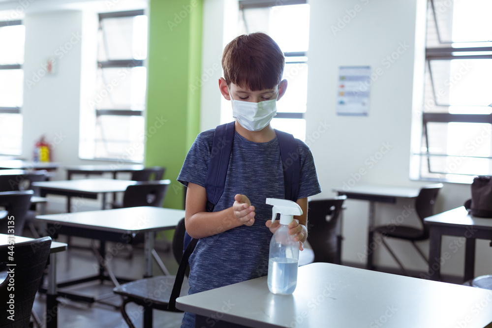 Fototapeta premium Caucasian schoolboy in classroom wearing face mask and schoolbag disinfecting hands