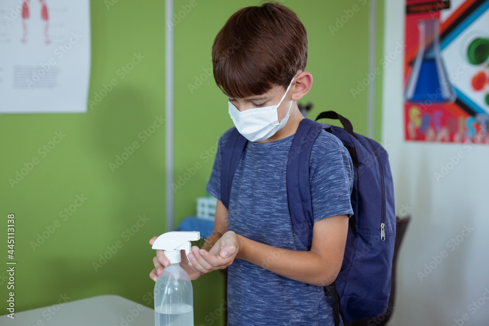 Fototapeta premium Caucasian schoolboy in classroom wearing face mask and schoolbag disinfecting hands