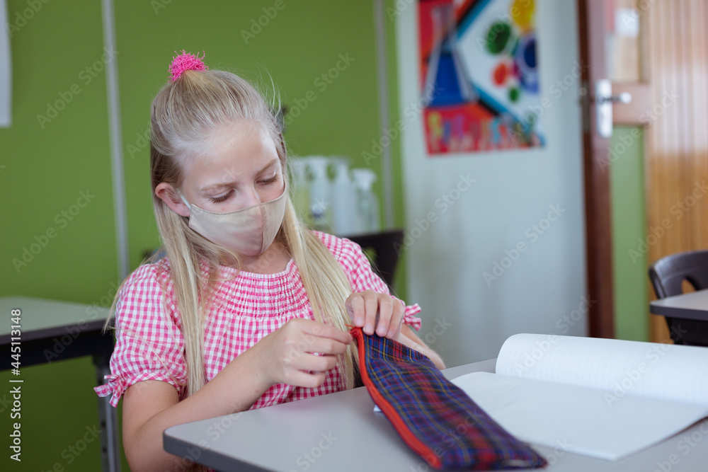 Fototapeta premium Caucasian schoolgirl sitting at desk in classroom wearing face mask