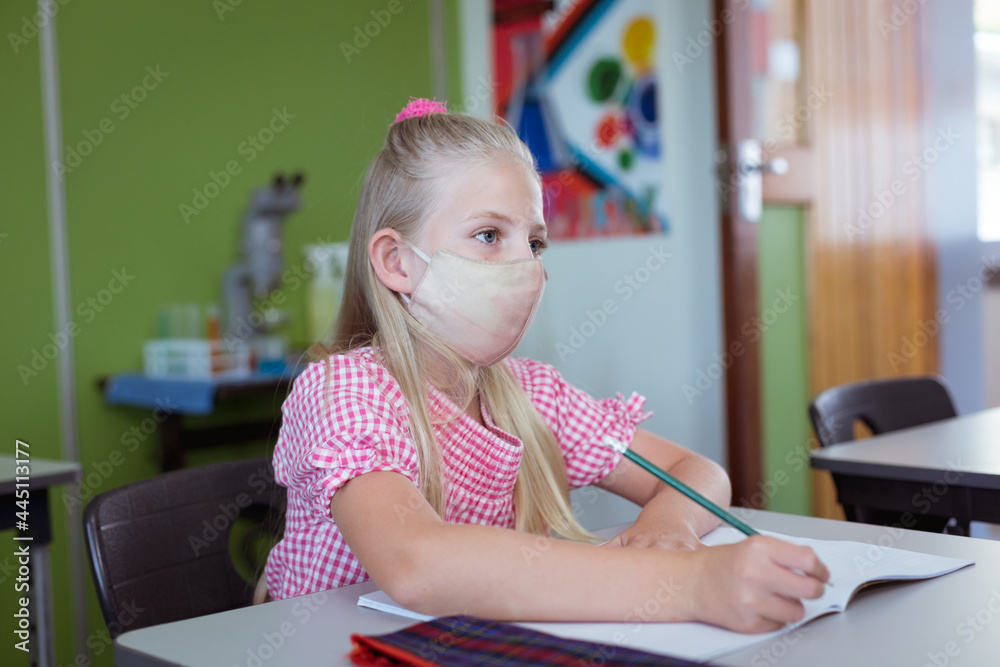 Fototapeta premium Caucasian schoolgirl sitting at desk in classroom wearing face mask during lesson