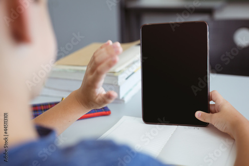 Caucasian schoolboy in classroom sitting at desk using tablet and waving, with copy space on screen