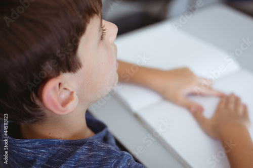 Blind caucasian schoolboy sitting at desk reading braille book with fingers