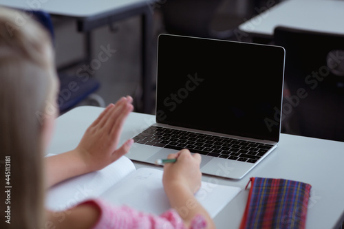 Caucasian schoolgirl at desk in classroom waving and using laptop, with copy space on screen