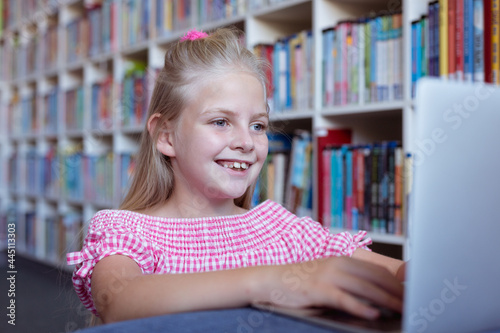 Smiling caucasian schoolgirl at desk in school library using laptop