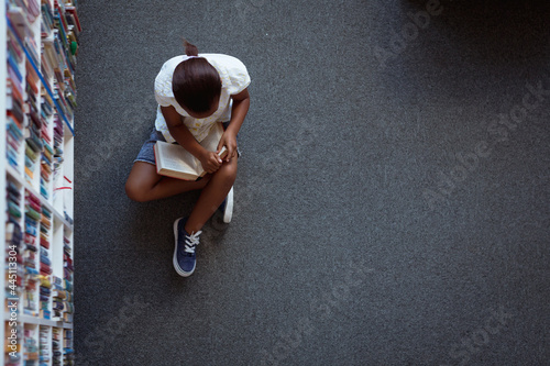 Overhead view of african american schoolgirl sitting on floor reading book in school library