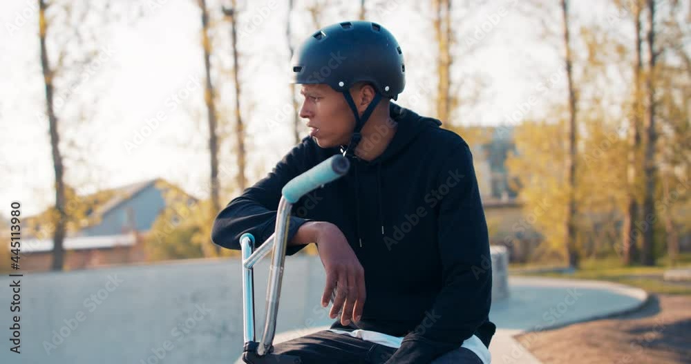 Young boy with dark complexion, sitting on bike leaning against handlebars, helmet on head, sitting in park on ramp, looking around, waiting for someone, friends Young boy with dark complexion, sitting on bike leaning against handlebars, helmet on head, sitting in park on ramp, looking around, waiting for someone, friends