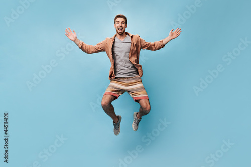 Joyful trendy man in brown shirt, grey t-shirt, shorts and sneakers looking into camera and jumping on isolated background..