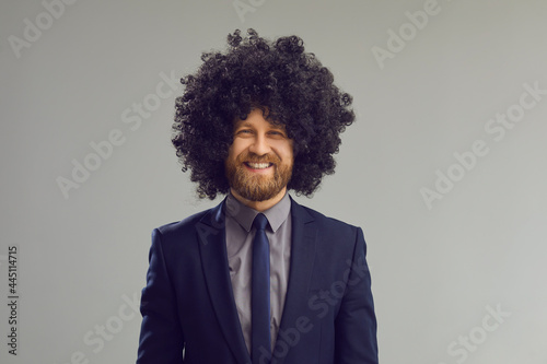 Headshot of cheerful smiling young man with crazy curly hair. Portrait of happy positive stylish bearded businessman in suit, necktie and funny wig looking at camera standing on grey studio background