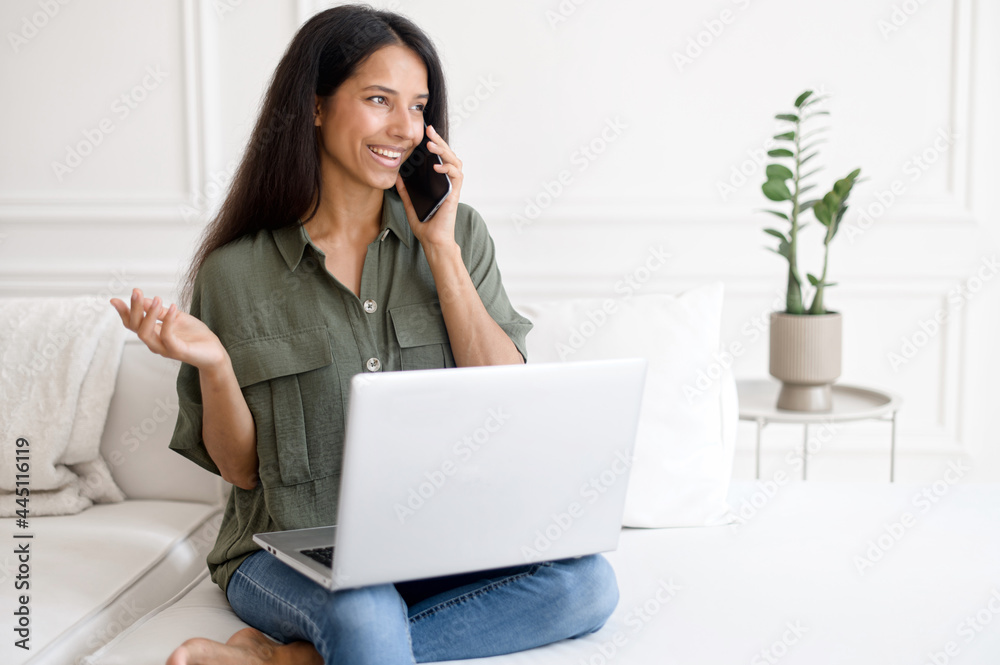 Indian woman with laptop talking on mobile phone sitting on the comfortable couch at home. Mixed-race female freelancer talking on the smartphone. Student girl using laptop during phone conversation Indian woman with laptop talking on mobile phone sitting on the comfortable couch at home. Mixed-race female freelancer talking on the smartphone. Student girl using laptop during phone conversation